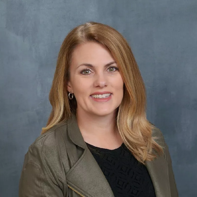 A professional woman is pictured wearing an olive green jacket over a black top. The setting is a neutral gray studio background, emphasizing a formal and polished appearance. Her blonde hair is styled neatly, complementing the overall professional tone of the image.